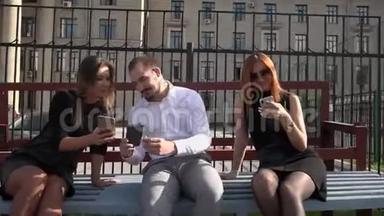 两名女<strong>学生</strong>和一名<strong>男学生</strong>在阳光明媚的天气里坐在长凳上用智能手机